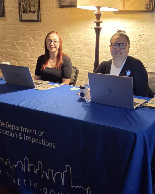 A person with long red hair and a person with glasses sit at a table with two laptops. The table is covered with a blue cloth displaying the "Seattle Department of Construction & Inspections" logo. They are in a room with a lamp and framed pictures on the brick wall behind them.