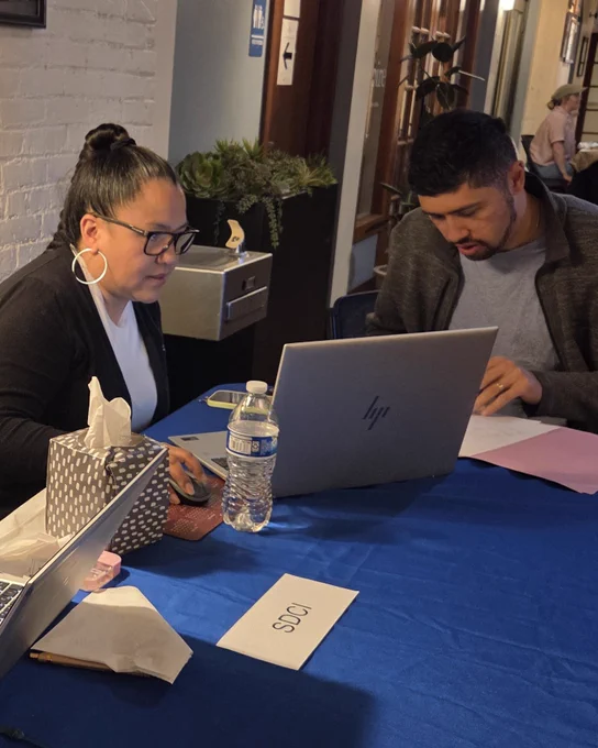Two people seated at a table working on laptops. One is writing on paper, the other is looking at a laptop screen. Items on the table include tissues, a water bottle, papers, and a name card labeled "SDCI"