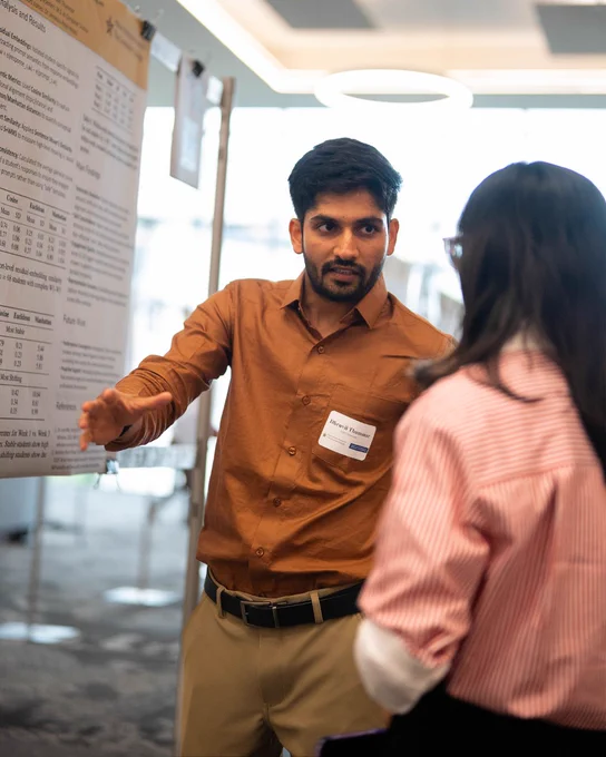 Man in brown shirt explaining research poster to woman in striped blouse at conference.