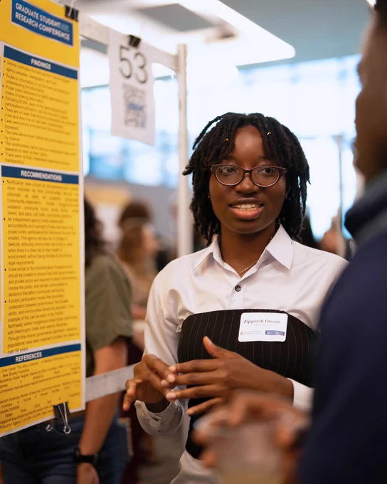 Graduate student presenting research findings at a conference poster session, engaging with an attendee.