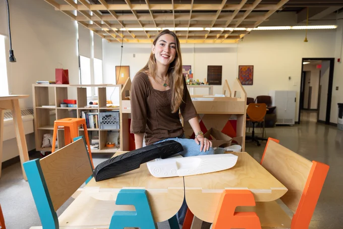 Person smiles while sitting on a table. The background features a studio setting.