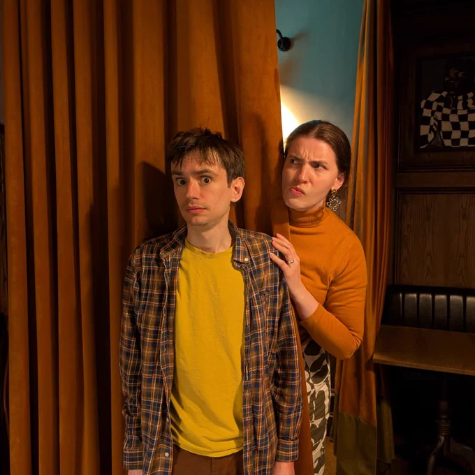 Photograph. Two people stand by or behind a set of wall length curtains. A man in front of the curtain looks to camera and appears to be slightly scared or intimidated. A woman, who stands behind the curtain, has a hand on his shoulder, but looks to the side, eyebrows furrowed and pursed lips.
