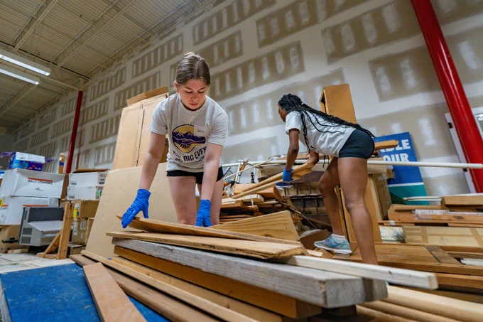 A wide photo of two students wearing blue gloves organizing wooden planks.
