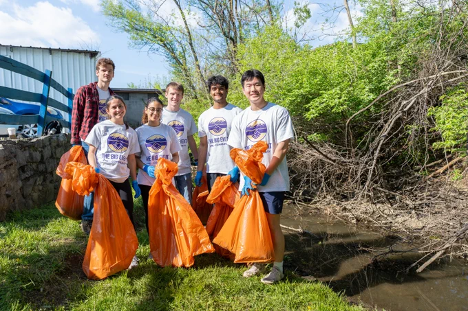 A wide photo of six students wearing “The Big Event” T-shirts hold orange trash bags and wear blue gloves. They are standing next to a trees and a body of water.
