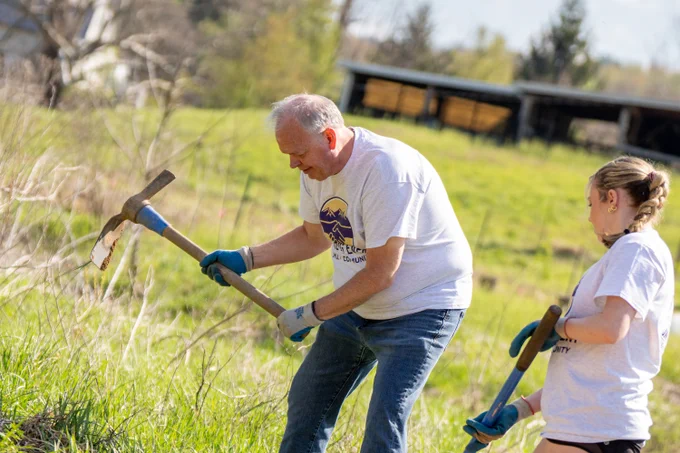 A wide photo of President Jim using a tool to dig into dirt in an open field, while a female student stands behind him holding another tool. 
