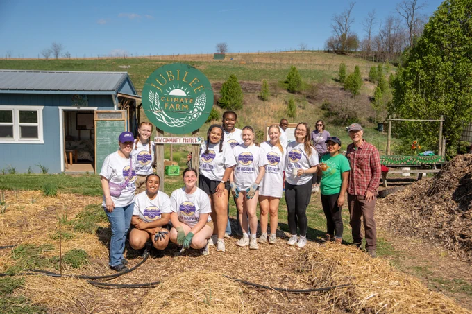 A wide photo of a group of students and faculty smiling in front of a Jubilee Climate Farm sign with fields and trees in the background.
