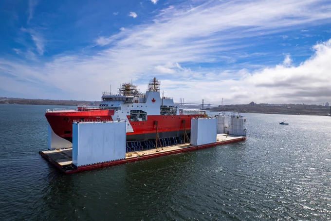 A red Canadian Coast Guard vessel is moored on a body of water, with large light‑coloured rectangular panels along its sides. The shoreline and a partly cloudy sky are visible in the background.