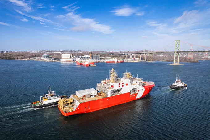 A red Canadian Coast Guard vessel sails on a waterway accompanied by two tugboats. A bridge and urban buildings are visible in the background under a lightly clouded blue sky.