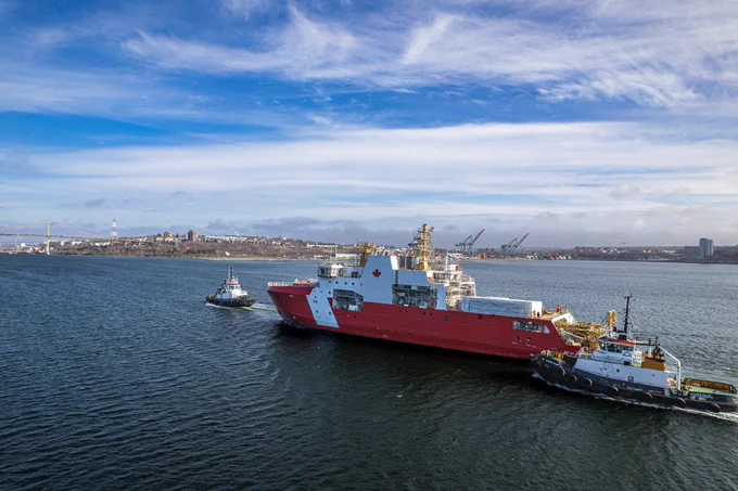 A large red Canadian Coast Guard vessel sails on a waterway, accompanied by two tugboats, with a city visible in the background under a partly cloudy sky.