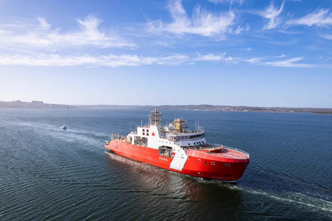 A red Canadian Coast Guard vessel sails across open water, viewed from above, leaving a wake behind it. The shoreline is visible on the horizon under a lightly clouded blue sky.