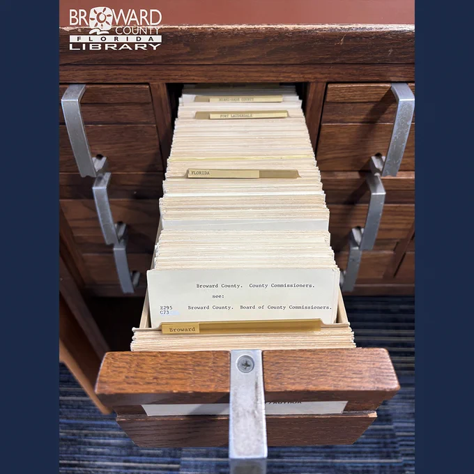 A wooden card catalog drawer is open, displaying index cards with typed labels. The visible card reads "Broward County. County Commissioners," suggesting a library reference system. Metal handles are visible on the cabinet, and the logo "Broward County Florida Library" is printed at the top of the image.
