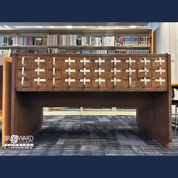 A wooden library card catalog with numerous small drawers, each featuring a metal handle and a paper label. It's situated under bookshelves filled with a variety of books. A logo for Broward County Library, Florida, is visible in the bottom left corner.