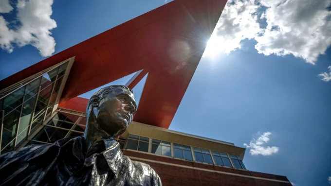 Bronze statue of a figure in the foreground, shot from a low angle against a bright blue sky with scattered clouds, in front of a modern building featuring a dramatic angular red architectural canopy with sun flare visible at its apex.