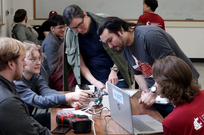 Group of students gathered around a table working on an electronics project, with a laptop, wires, and hardware components spread out, during an IEEE hardware hackathon in a classroom setting.