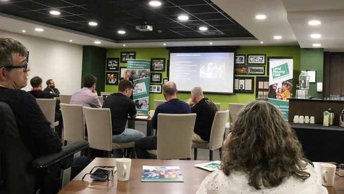 A member of the Give Vision team, presenting next to a projector screen, in front of a room of people seated around tables.
