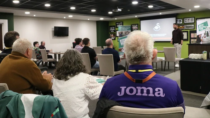 Swansea City's CEO, presenting next to a projector screen, in front of a room of people seated around tables.