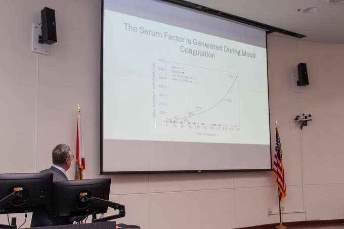 Gabor Tigyi looks up at a screen and presents the content. The screen features headline text that reads "The Serum Factor is Generated During Blood Coagulation" along with a graph.