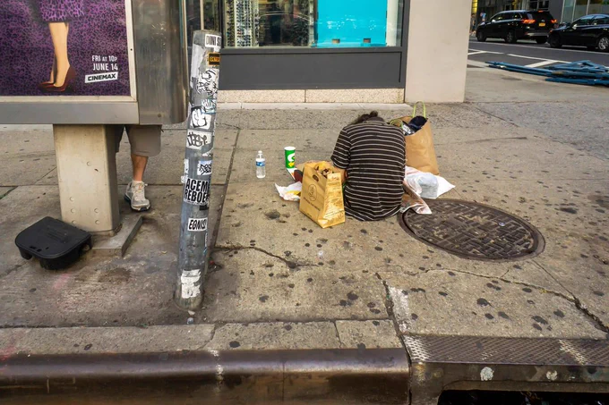 A person sits on a city sidewalk near a phone booth and a utility pole covered in stickers. The person is surrounded by a few paper bags and a drink cup. The environment is urban, with cars parked on the street nearby.