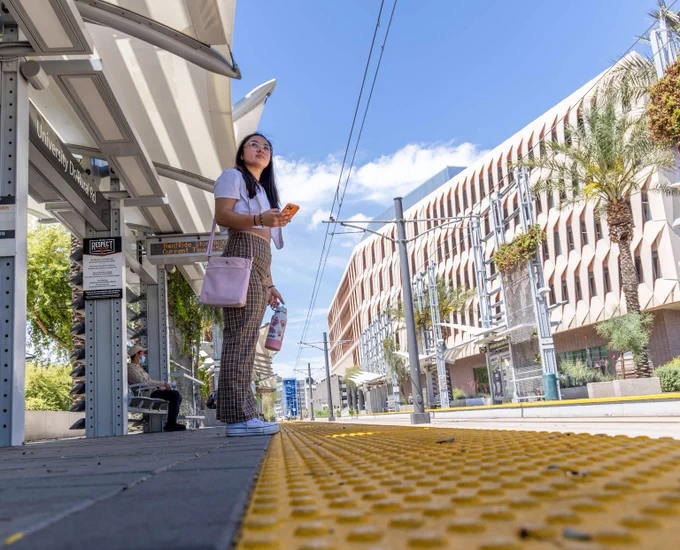 Woman standing on a sunny urban tram platform holding a water bottle and phone, with modern buildings and palm trees in the background.
