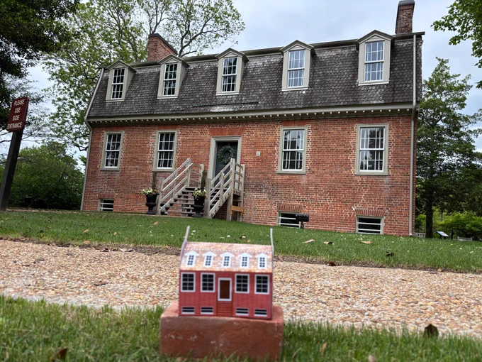 Small cardstock model of the Francis Land House sitting on the ground in front of the actual Francis Land House, a historic brick home in Virginia Beach.