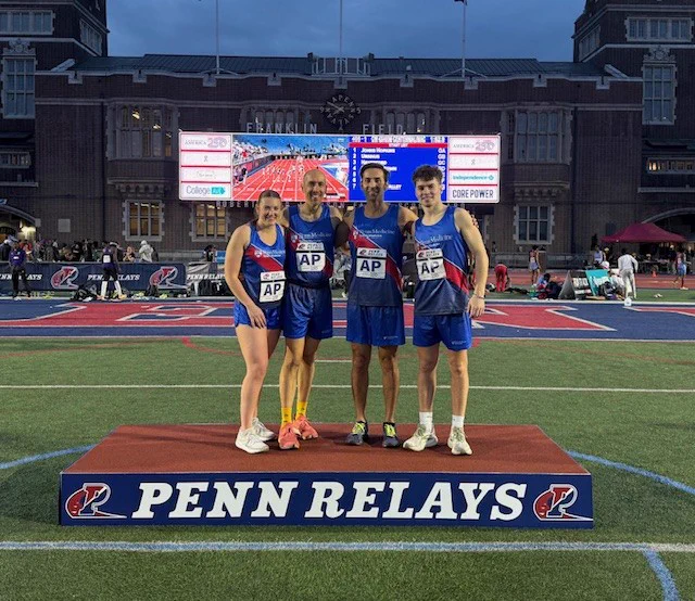 Four runners in blue uniforms, two women and two men, stand on a \"PENN RELAYS\" podium. They are on a green field in front of Franklin Field stadium, under a scoreboard showing \"AMERICA text@1\".