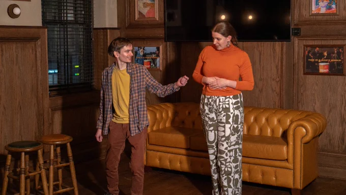 Photograph. Two people onstage. Both are standing. One has a knee bent and one arm out. The second person looks at the other's posture, keen to learn.