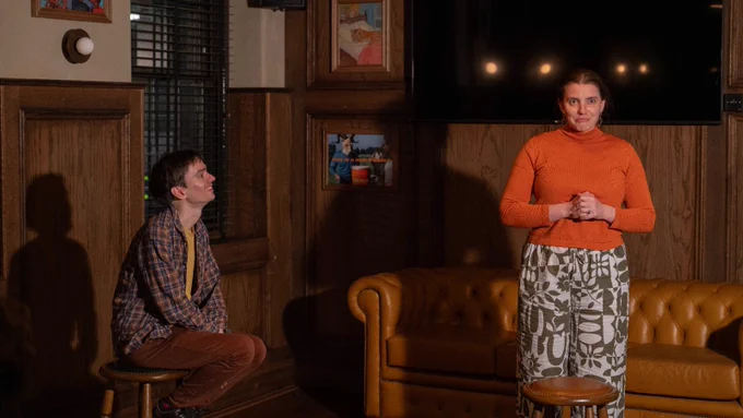 Photograph. Two people onstage. One sits on a stool and faces to the right. They look fondly at a second person. A second person stands onstage, looking forward, with their hands clasped together in front of their chest.