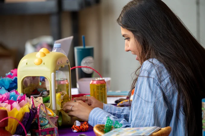 Person with long hair playing a small yellow claw machine on a colorful table filled with toys and snacks.