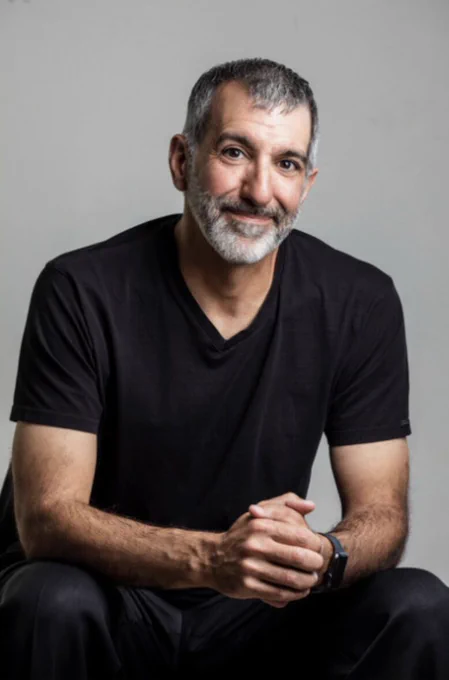 A man wearing a black shirt and pants sits in front of a grey photo backdrop.