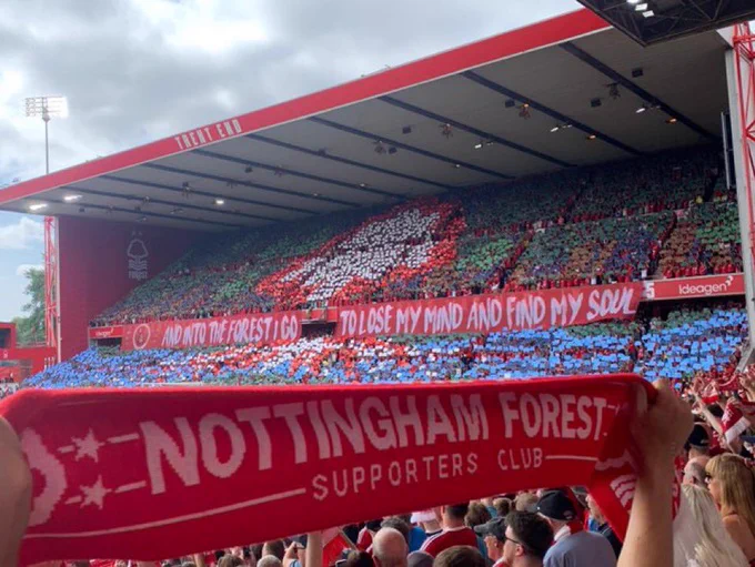 Photo of a packed Trent End stand at Nottingham Forest’s City Ground Stadium. The image is facing diagonally right from within the Brian Clough stand and at the foot of the picture, taking up the bottom third is a red scarf with Nottingham Forest written in white from left to right. There is a colourful display shown in the Trent End stand with the top of the Forest crest (a tree) depicted in the upper tier of the stand. The bottom tier is predominantly blue whilst a banner stretches the width of the stand directly between both tiers, the wording on this banner is as follows: “And into the Forest I go, to lose my mind and find my soul!”