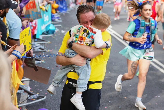 Man in yellow hugging a small child at a marathon finish line while runners pass and spectators cheer.
