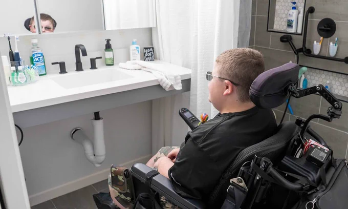 A person in a powerchair is facing a bathroom sink and mirror, with hygiene products on the counter. Reflection visible in the mirror.