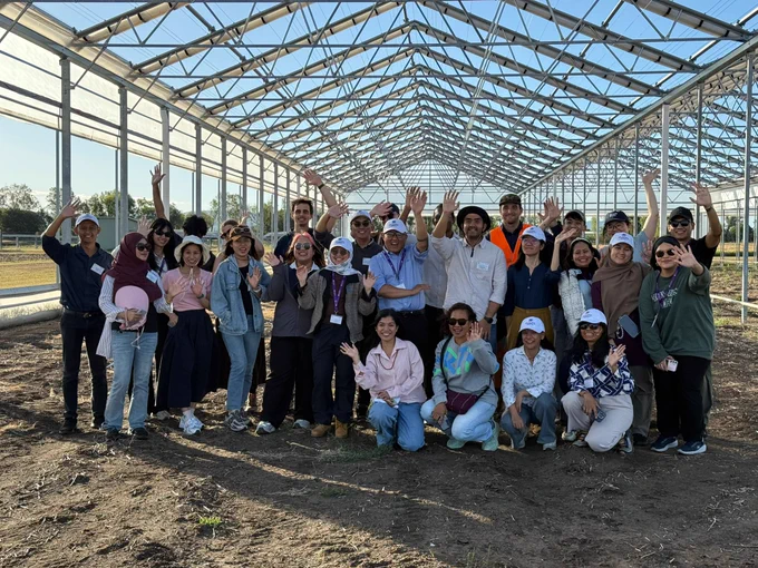 Group smiling and waving inside a large metal greenhouse frame under clear blue sky.