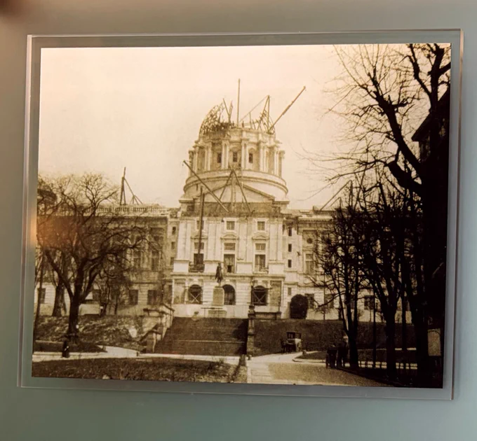 Sepia-toned photo of a large, ornate building under construction, with scaffolding on its domed roof. Leafless trees frame the scene. The photo is in a glass casing against a light green background.