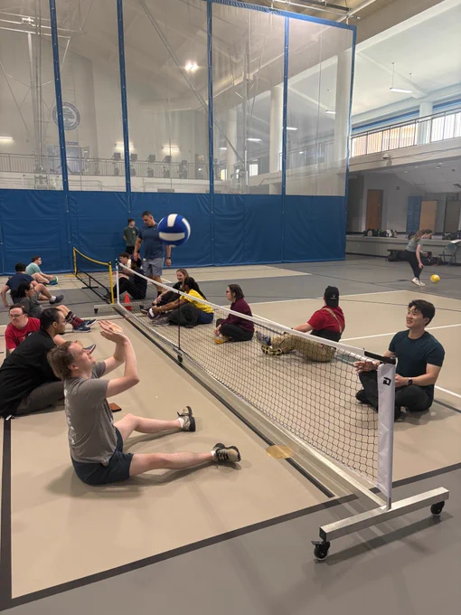 A back angle photo of individuals on a gymnasium floor playing sitting volleyball. A volleyball is in the air and a net is stationed between many people on either side.