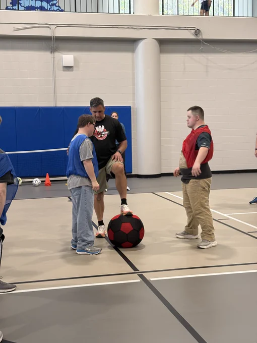 A photo primarily focused on three individuals standing and playing futsal. One individual is talking to another with his foot up on a large soccer ball. Other individuals are gathered around to play just out of of the shot.