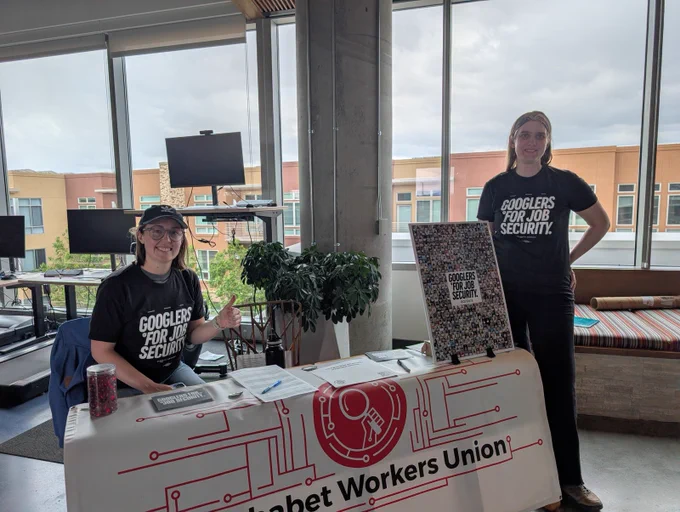 two AWU members display a large poster collage of Googlers for Job Security supporters, wearing Googlers for Job Security t-shirts and tabling in a cafeteria on a Colorado Google campus with an Alphabet Workers Union banner draped over the table. 