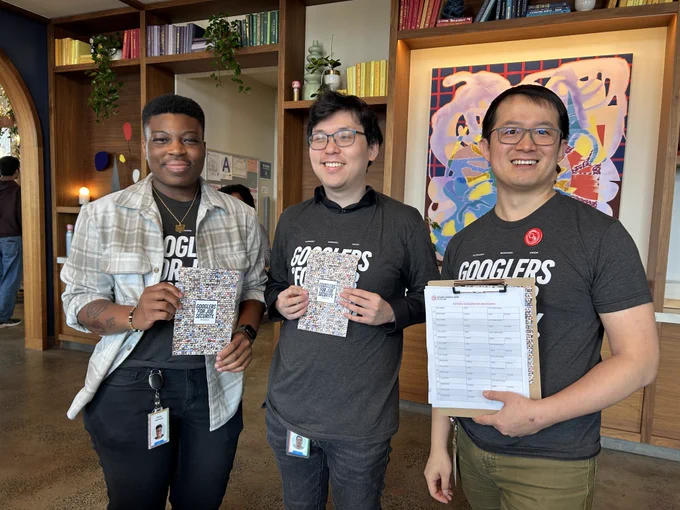 Three AWU members wearing Googlers for Job Security t-shirts in a Google cafeteria in NYC holding clipboards and flyers with a big collage of Googlers for Job Security supporters. 