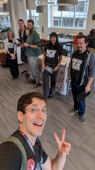 A group selfie of six AWU members tabling at a Google campus in Pittsburgh with a big banner of photos of Googlers for Job Security supporters, holding clipboards and flyers.