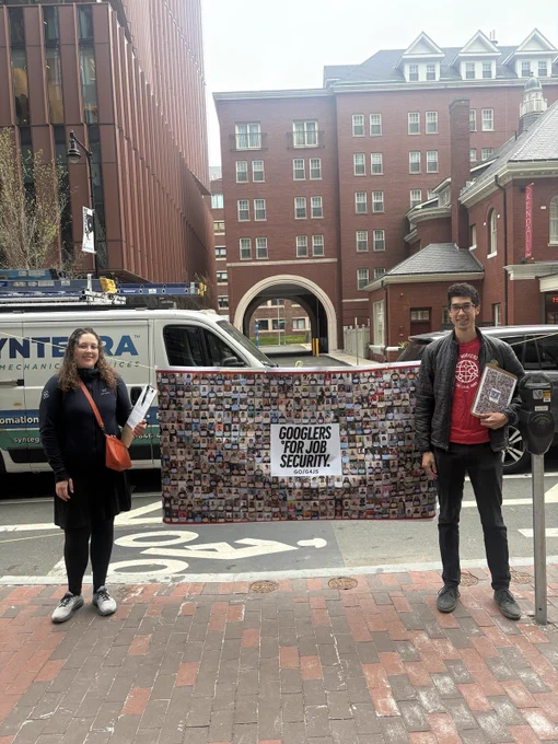 two AWU members pose outside a Google campus in Cambridge with a giant banner with a collage of photos of Googlers for Job Security supporters, holding clipboards and flyers.