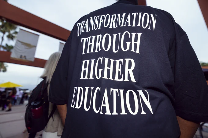 Person wearing a T-shirt with the phrase "Transformation Through Higher Education" written in bold letters on the back. The setting appears to be an outdoor event with visible structures in the background.