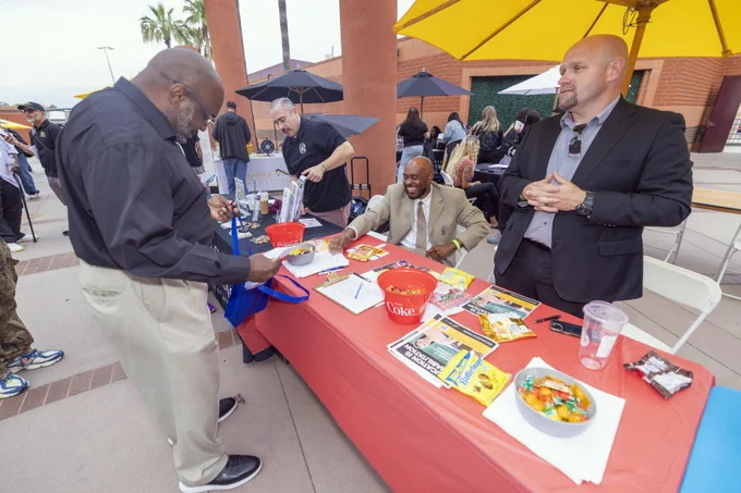 People are gathered around a table at an outdoor event. The table is covered with a red cloth and has brochures, snacks, and a bucket. Two individuals are seated at the table, and one stands, engaging with a visitor. Yellow umbrellas provide shade in the background.
