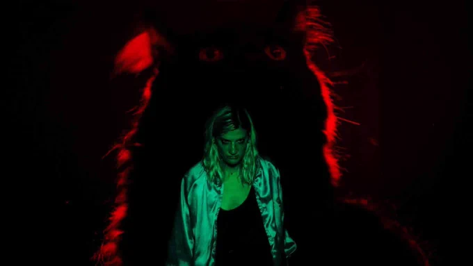 Still from the film 'Booger' of an individual in green lighting and a large cat lit in red, looming behind her.