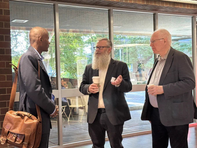 Three people are engaged in conversation by large windows. Two are holding coffee cups. They appear to be at a casual or professional gathering.