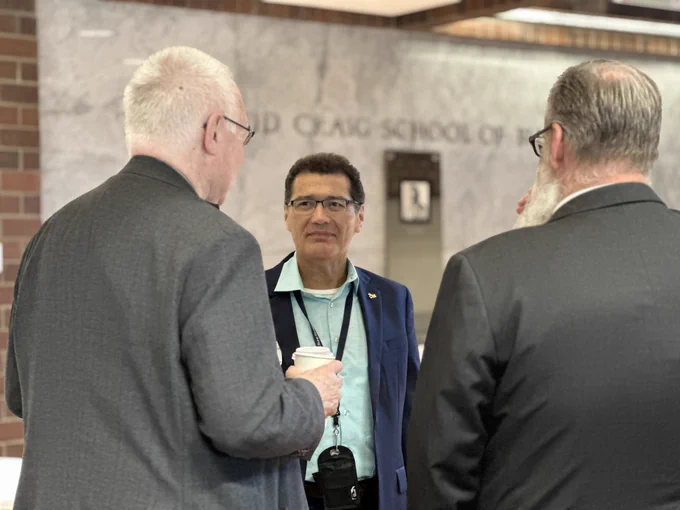 There are three individuals in conversation in front of a wall. One is holding a coffee cup and wearing a lanyard.