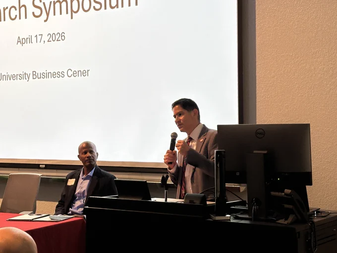 A person at a podium speaks into a microphone, while another is seated at a table, listening. The screen behind them displays "Research Symposium, April 17, 2026, University Business Center."
