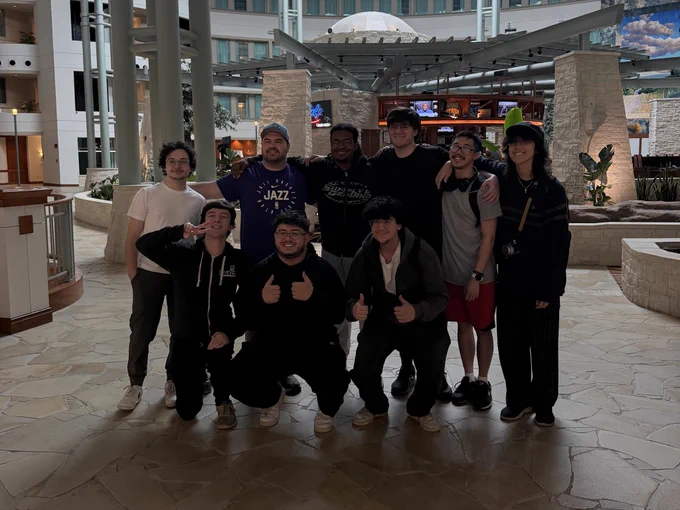 A group of nine people posing together in a hotel lobby. The space features stone flooring, tall columns, and a mix of natural and modern design elements — including plants, stone walls, and a restaurant or café visible in the background with a neon sign. Six people stand in the back while four crouch in front of them, making peace signs or thumbs-up gestures.