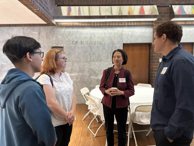 A group of four people are standing in a circle, engaging in conversation. They are in an indoor setting with a backdrop that includes white chairs and round tables. The wall in the background has the text "School of Business."