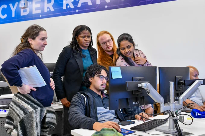 A group of five people collaborating around computer monitors in a modern office with a Cyber Range banner overhead.