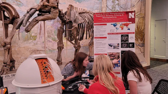 Bridget Slagle points to data on a large sign while talking with two others with sensors on their fingers to learn about the Galileo Tactile Stimulus System; mammoth skeleton stands in the background